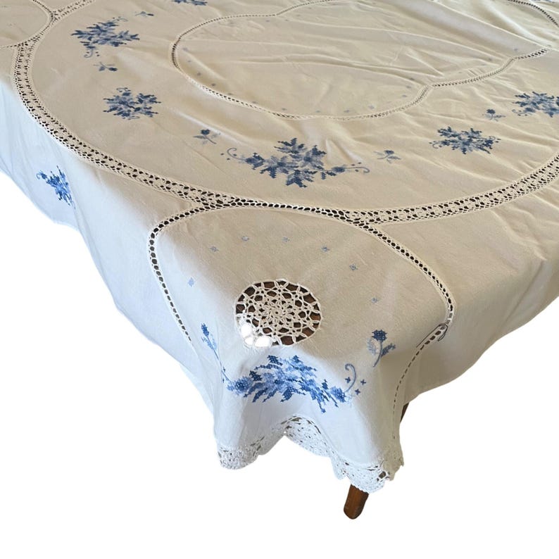 Puede incluir: Mantel blanco con bordado floral azul y ribete de encaje de ganchillo. El mantel presenta un dise&ntilde;o circular con flores bordadas y detalles de encaje calado. Una cubierta de mesa de estilo vintage.