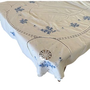 Puede incluir: Mantel blanco con bordado floral azul y ribete de encaje de ganchillo. El mantel presenta un dise&ntilde;o circular con flores bordadas y un delicado borde de encaje calado. Una esquina tiene un dise&ntilde;o circular de ganchillo.