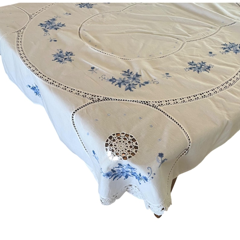 Puede incluir: Mantel blanco con bordado floral azul y ribete de encaje de ganchillo. El mantel presenta un dise&ntilde;o circular con flores bordadas y un delicado borde de encaje calado. Una esquina tiene un dise&ntilde;o circular de ganchillo.