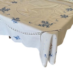 Puede incluir: Mantel blanco con bordado floral azul y borde de encaje festoneado. El mantel tiene un centro redondo y est&aacute; drapeado sobre una mesa, mostrando el detallado trabajo de aguja y el dise&ntilde;o.