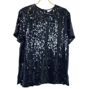 Puede incluir: Una blusa negra de lentejuelas con mangas cortas y cuello redondo. La blusa está hecha de una tela brillante y brillante.