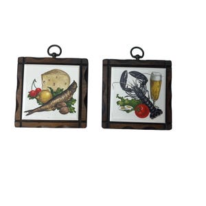 Peut inclure: Deux tableaux carrés avec des cadres en bois foncé. L'un représente du fromage, des cerises, un citron, un poisson et des noix. L'autre montre un homard, de la bière, une tomate et des œufs. Chacun a une boucle en métal pour accrocher.