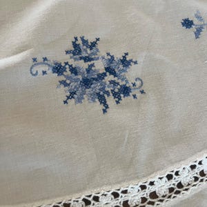 Puede incluir: Mantel de lino color crema con bordado floral de punto de cruz azul. El dise&ntilde;o presenta un gran motivo floral y peque&ntilde;os acentos. Un borde de encaje de ganchillo blanco adorna el borde, agregando un toque delicado al textil vintage.