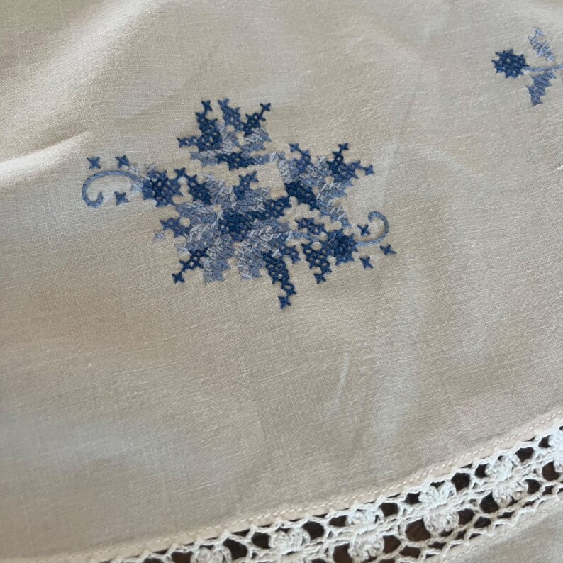 Puede incluir: Mantel de lino color crema con bordado floral de punto de cruz azul. El dise&ntilde;o presenta un gran motivo floral y peque&ntilde;os acentos. Un borde de encaje de ganchillo blanco adorna el borde, agregando un toque delicado al textil vintage.