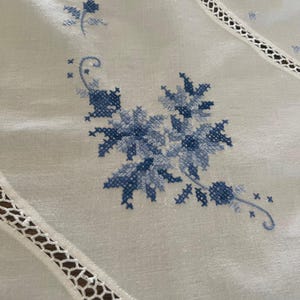 Puede incluir: Primer plano de un mantel blanco con bordado floral azul a punto de cruz. El dise&ntilde;o presenta flores y hojas estilizadas. Los bordes del mantel tienen un delicado ribete de encaje calado.