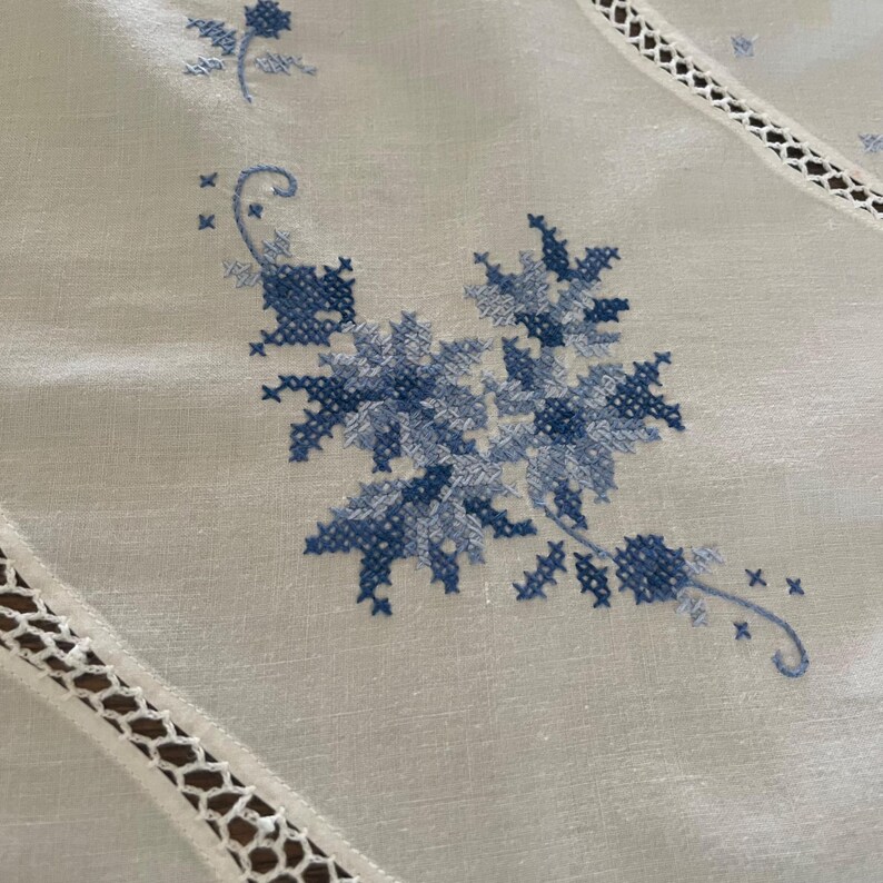 Puede incluir: Primer plano de un mantel blanco con bordado floral azul a punto de cruz. El dise&ntilde;o presenta flores y hojas estilizadas. Los bordes del mantel tienen un delicado ribete de encaje calado.