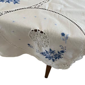 Puede incluir: Mantel crema con bordado floral azul y borde de encaje de ganchillo. El mantel presenta un dise&ntilde;o circular de ganchillo y est&aacute; drapeado sobre una mesa de madera. Se ve una sola pata.