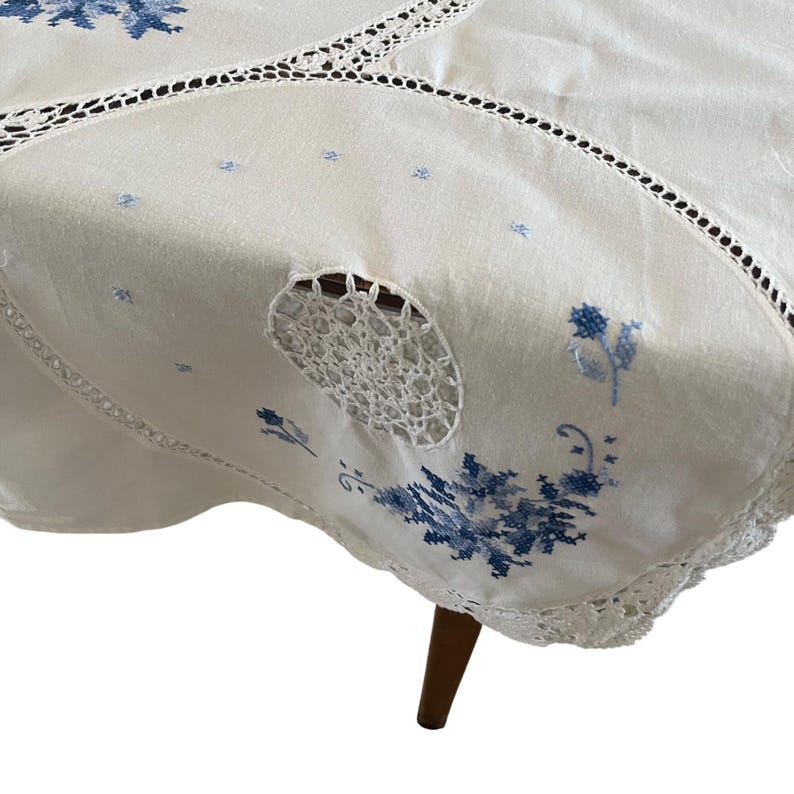 Puede incluir: Mantel crema con bordado floral azul y borde de encaje de ganchillo. El mantel presenta un dise&ntilde;o circular de ganchillo y est&aacute; drapeado sobre una mesa de madera. Se ve una sola pata.