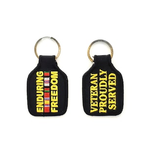 May include: Two black keychains with yellow embroidered text. One keychain says "Enduring Freedom" with a small embroidered American flag. The other keychain says "Veteran Proudly Served".