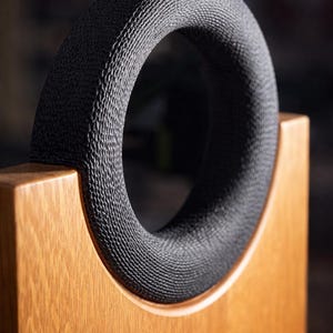 May include: A close-up of a speaker with a black, textured ring and a wooden frame. The ring is circular and has a woven pattern. The wooden frame is light brown and has a natural wood grain.