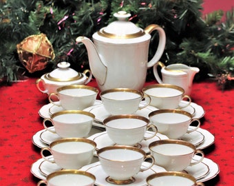 May include: A white porcelain tea set with gold trim, including a teapot, sugar bowl, creamer, and multiple teacups and saucers. The set is arranged on a red tablecloth, with Christmas decorations in the background.