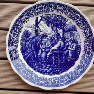 May include: A white and blue decorative plate with a scalloped edge. The plate features a detailed blue illustration of people gathered outdoors, with a floral border design. The plate is likely ceramic.