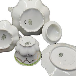 May include: A white porcelain tea set with a scalloped edge design. The set includes a teapot, sugar bowl, creamer, teacup, and saucer. The pieces have a gold trim and a decorative pattern on the bottom. The set is arranged on a white surface.