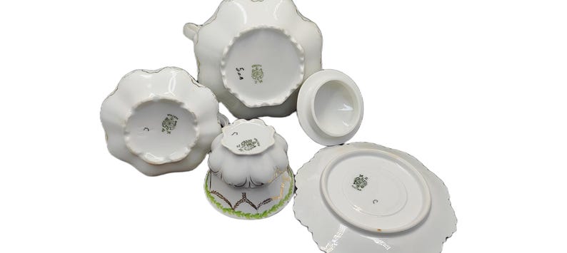 May include: A white porcelain tea set with a scalloped edge design. The set includes a teapot, sugar bowl, creamer, teacup, and saucer. The pieces have a gold trim and a decorative pattern on the bottom. The set is arranged on a white surface.