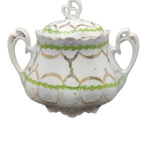 May include: White porcelain sugar bowl with lid, featuring gold and green decorative patterns. The bowl has two handles and a decorative finial on the lid. A classic piece for serving sugar.