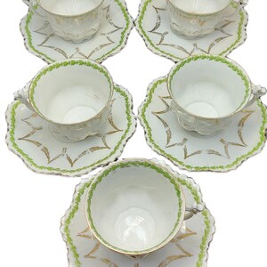 May include: Set of five white teacups and saucers. Each cup and saucer features a scalloped edge with green and gold leaf accents. The cups have a delicate, embossed design.