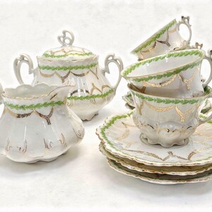 May include: A vintage porcelain tea set with a white base, adorned with green and gold decorative accents. The set includes teacups, saucers, a creamer, and a sugar bowl with a lid. The teacups are stacked, showcasing the intricate design.