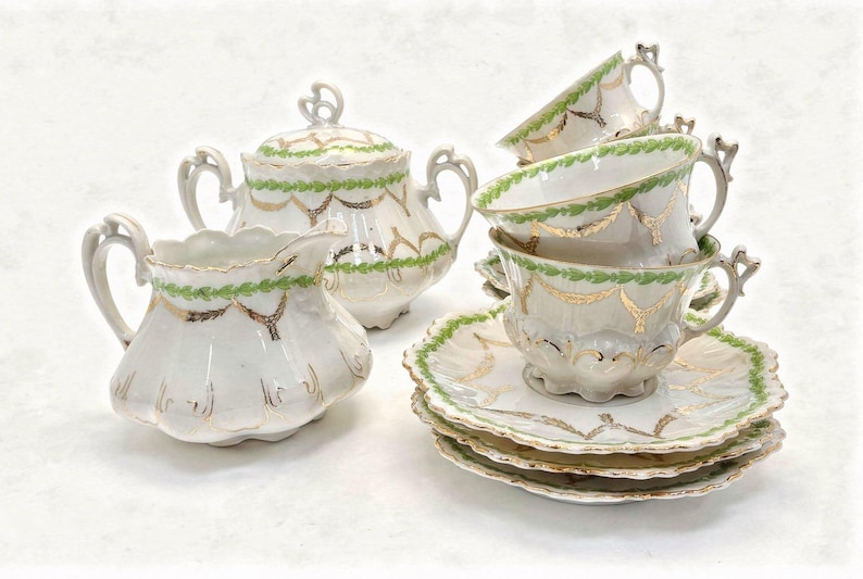 May include: A vintage porcelain tea set with a white base, adorned with green and gold decorative accents. The set includes teacups, saucers, a creamer, and a sugar bowl with a lid. The teacups are stacked, showcasing the intricate design.