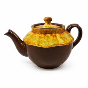 Op de afbeelding: Een keramische theepot met een donkerbruine basis en een contrasterend geel en oranje druipglazuur op het bovenste gedeelte en het deksel. De theepot heeft een gebogen tuit, een afgerond handvat en een kleine knop op het afneembare deksel.