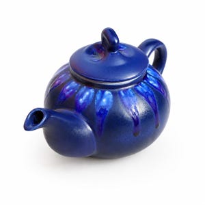 May include: A round ceramic teapot in a deep cobalt blue glaze featuring a decorative pattern of lighter blue, petal-like drips radiating from the lid down the upper body. The teapot includes a matching lid with a central handle and a curved spout.
