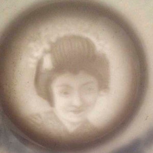 May include: A close-up of a ceramic plate featuring a raised image of a woman with a traditional Japanese hairstyle. The image is in shades of grey and white.