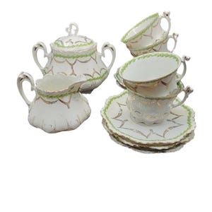 May include: A white porcelain tea set with gold trim and green accents. The set includes teacups, saucers, a sugar bowl with a lid, and a creamer. The tea set has a delicate, vintage design.