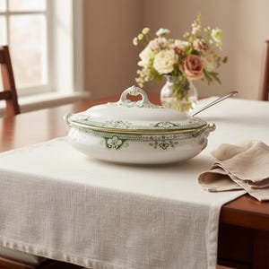 Può includere: Una zuppiera in ceramica bianca con coperchio, con un motivo floreale verde e bordatura dorata. Il piatto ovale è posto su una tovaglia bianca. Un cucchiaio d'argento è appoggiato sul tavolo e un vaso di fiori è sullo sfondo.