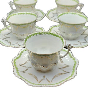 May include: Set of six white teacups and saucers with green and gold leaf design. The teacups have ornate handles and the saucers have a scalloped edge. The teacups and saucers are arranged on a white background.
