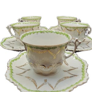 May include: A set of six antique teacups and saucers. The cups and saucers are white with gold and green leaf designs. The saucers have a scalloped edge.