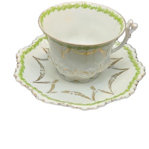 May include: A vintage teacup and saucer set. The cup and saucer are white with gold accents and a green leaf design around the rim and edges. The cup has a decorative handle.