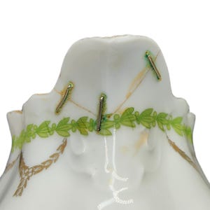 May include: Close-up of a white porcelain object with gold and green decorative accents. The object has a scalloped edge and is adorned with gold leaf patterns and a green leaf garland. Metal staples are visible.