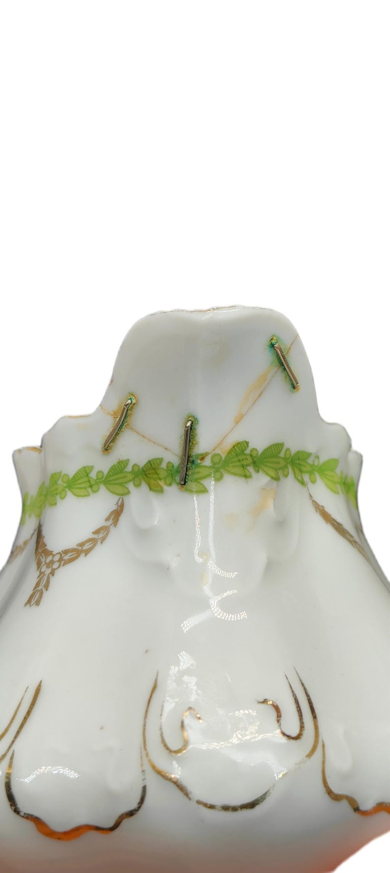 May include: Close-up of a white porcelain object with gold and green decorative accents. The object has a scalloped edge and is adorned with gold leaf patterns and a green leaf garland. Metal staples are visible.