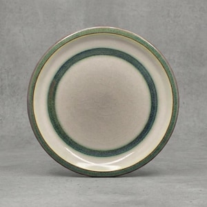 Può includere: Un piatto rotondo in ceramica con un centro color crema e un bordo verde e turchese. Il piatto ha un design semplice ed elegante, adatto per servire cibo. Il piatto è probabilmente per l'uso quotidiano.