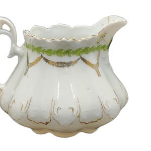 May include: White porcelain creamer with a scalloped rim and a decorative handle. The creamer features gold accents and green leaf garland around the top. The creamer has a bulbous shape and a spout for pouring.