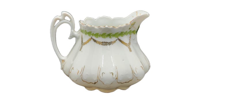 May include: White porcelain creamer with a scalloped rim and a decorative handle. The creamer features gold accents and green leaf garland around the top. The creamer has a bulbous shape and a spout for pouring.