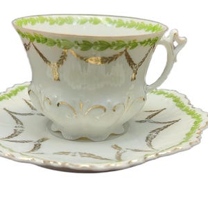 May include: A white teacup and saucer set with gold and green accents. The teacup has a scalloped rim with green leaves and gold garland designs. The saucer has a matching scalloped edge with green and gold leaf patterns.