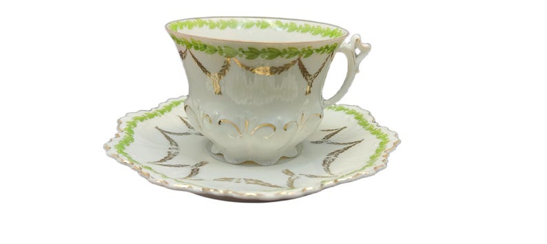 May include: A white teacup and saucer set with gold and green accents. The teacup has a scalloped rim with green leaves and gold garland designs. The saucer has a matching scalloped edge with green and gold leaf patterns.