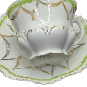 May include: White teacup and saucer set with green and gold decorative accents. The cup and saucer feature a scalloped edge and a repeating leaf and garland design. A classic tea set.