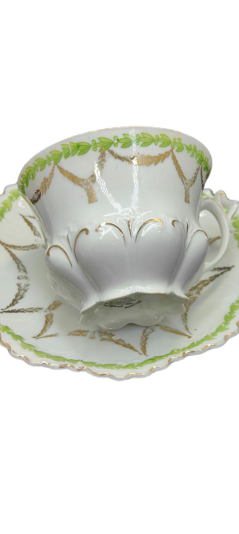 May include: White teacup and saucer set with green and gold decorative accents. The cup and saucer feature a scalloped edge and a repeating leaf and garland design. A classic tea set.
