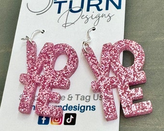 May include: Pink glitter "VOTE" earrings with silver hooks. The earrings are displayed on a white card with the text "HALF TURN Designs" and social media icons. The website address "halfturndesigns.com" is also visible.