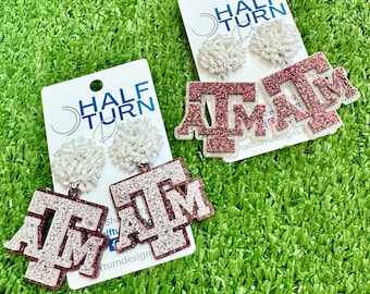 May include: A pair of earrings featuring a white pom-pom charm and a maroon glitter 'A&M' charm. The earrings are displayed on a white card with the text 'Half Turn Designs'.