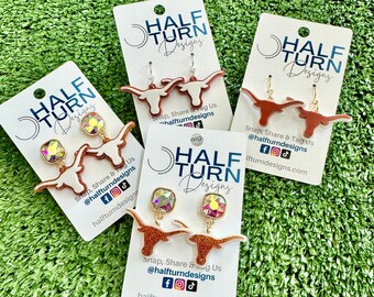 May include: Three pairs of earrings featuring a white and orange glitter Longhorn steer design. Each pair is displayed on a white card with the text "Half Turn Designs" and social media handles.