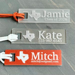May include: Three personalized luggage tags with a Texas state outline and a longhorn steer. The top tag is clear with the name "Jamie" and the text "Class of 2028". The middle tag is clear with the name "Kate" and the phone number "555-987-6543". The bottom tag is red with the name "Mitch" and the email address "mitch@notgmail.com".