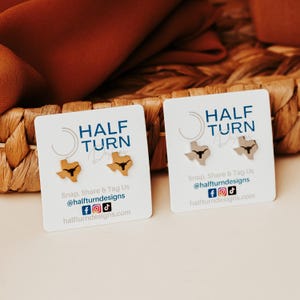 May include: Two pairs of stud earrings shaped like the state of Texas, each with a longhorn silhouette. One pair is gold-colored, the other is silver. The earrings are displayed on white cards with the text "HALF TURN Designs".