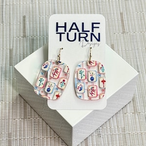 May include: A pair of clear acrylic earrings with gold-colored hooks. The earrings feature a rectangular shape with rounded corners and a colorful pattern of floral and geometric designs. The earrings are displayed on a white card with the text "HALF TURN Designs".