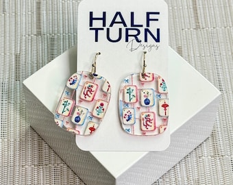 May include: A pair of clear acrylic earrings with gold-colored hooks. The earrings feature a rectangular shape with rounded corners and a colorful pattern of floral and geometric designs. The earrings are displayed on a white card with the text "HALF TURN Designs".