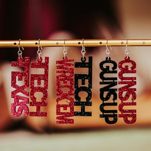 May include: Five pairs of glitter earrings with different words on them. The words are "Texel", "Tech", "Wrecked", "Guns Up", and "Guns Up". The earrings are red and black.