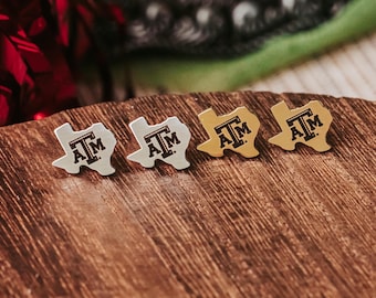 May include: Four Texas-shaped stud earrings. Two are silver-toned, and two are gold-toned. Each earring features the letters "A" and "M" in a bold, black font. The earrings are displayed on a wooden surface.