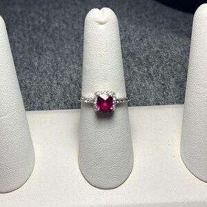 May include: A silver ring featuring a square, vibrant red gemstone, surrounded by a halo of smaller, clear stones. The ring is displayed on a white ring holder against a gray background.