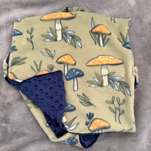 May include: A soft, reversible blanket featuring a whimsical mushroom and leaf pattern in shades of green, orange, and blue. The blanket's interior is a solid navy blue with a textured, dotted surface. The blanket is folded.